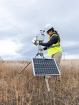 Field technician operating solar-powered environmental monitoring equipment in a grassland setting.