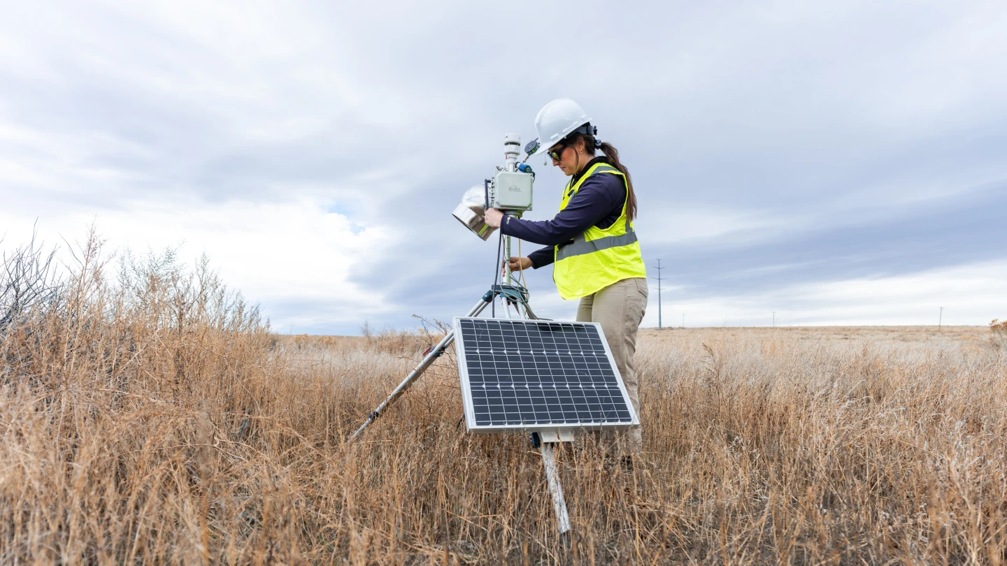 Field technician operating solar-powered environmental monitoring equipment in a grassland setting.