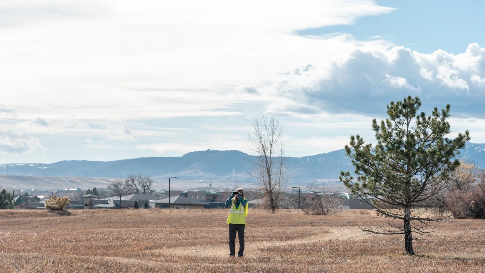 Woman wearing a yellow vest and looking through binoculars while standing in a field with homes and mountains in the background.