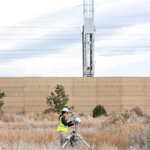 Technician in safety gear operating air quality monitoring equipment in a dry field near an industrial stack and power lines.