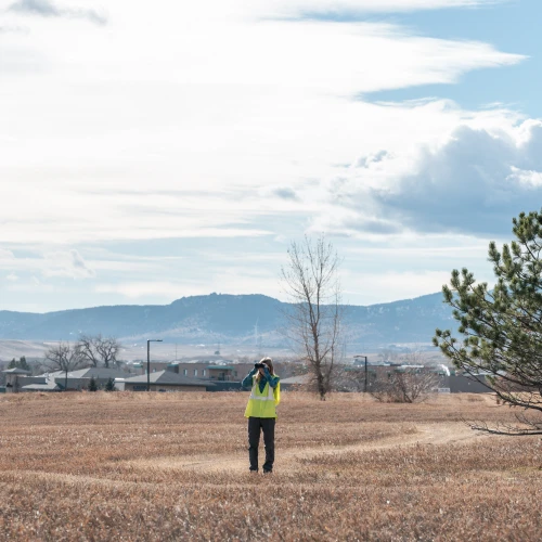 Woman wearing a yellow vest and looking through binoculars while standing in a field with homes and mountains in the background.