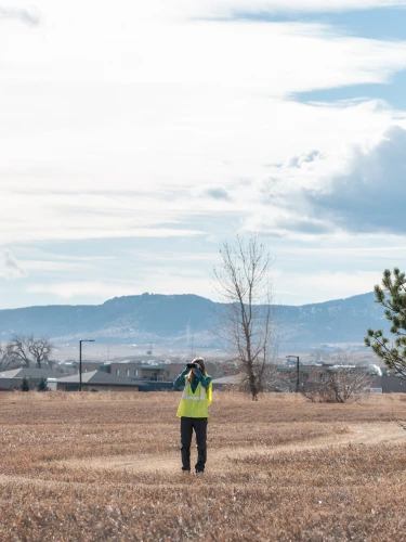 Woman wearing a yellow vest and looking through binoculars while standing in a field with homes and mountains in the background.