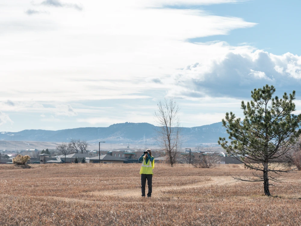 Woman wearing a yellow vest and looking through binoculars while standing in a field with homes and mountains in the background.