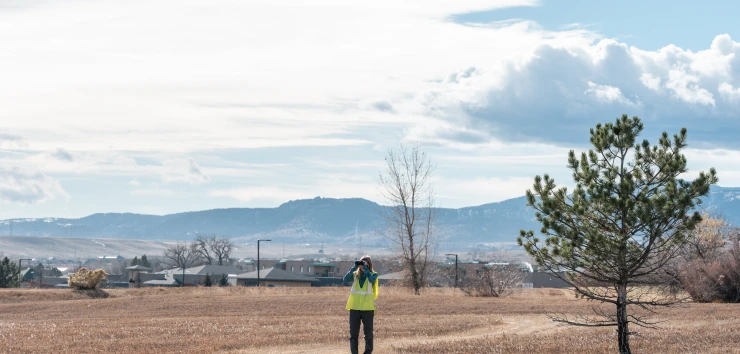 Woman wearing a yellow vest and looking through binoculars while standing in a field with homes and mountains in the background.