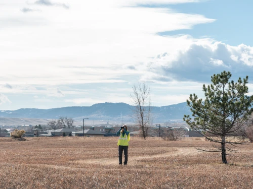 Woman wearing a yellow vest and looking through binoculars while standing in a field with homes and mountains in the background.