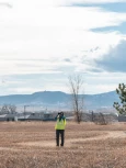 Woman wearing a yellow vest and looking through binoculars while standing in a field with homes and mountains in the background.